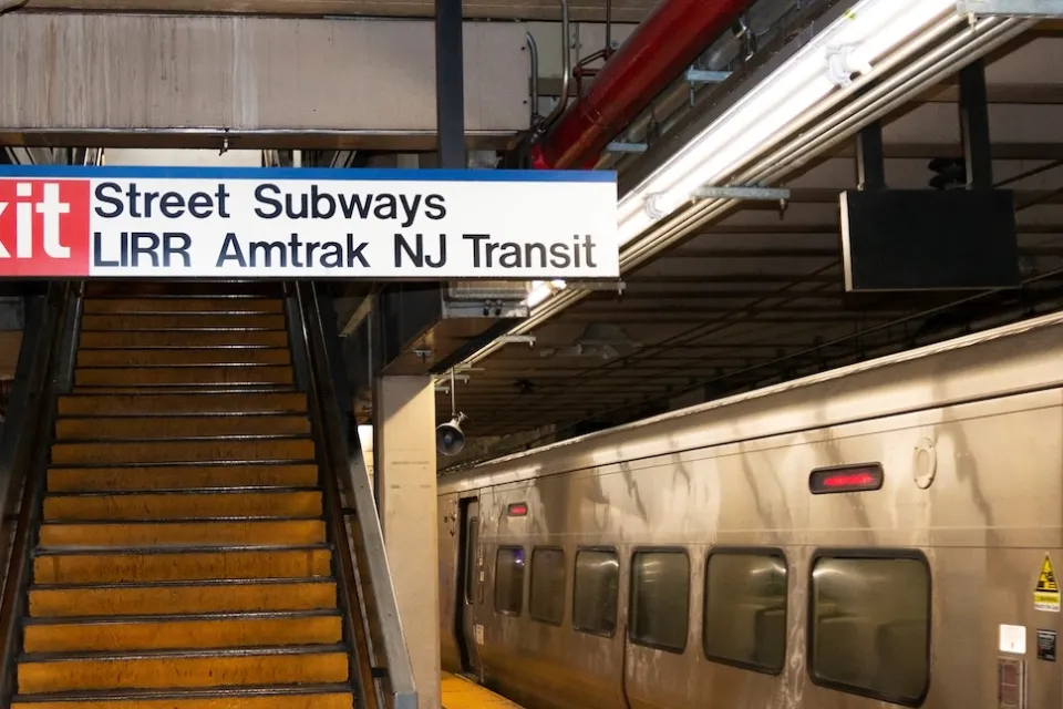 "Street Subway - LIRR - Amtrak - NJ Transit" with Exit sign in front of stairway on subway platform at Penn Station with silver train stopped at right.